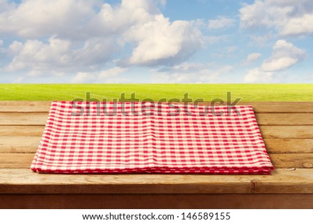 Empty wooden table with red checked tablecloth over beautiful landscape with green meadow and blue sky