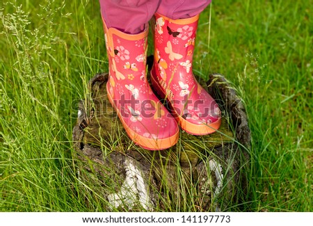 Child girl legs in pink galoshes standing on old tree stump with green grass around
