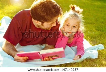 Father reading a book to his daughter while laying outdoor on the grass in the garden during sunset