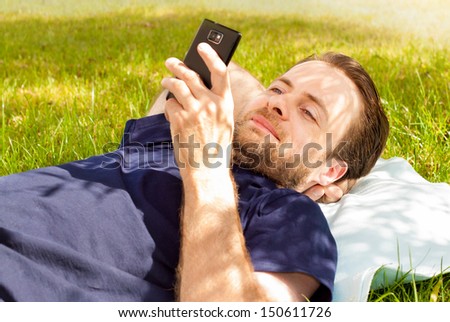 Happy Smiling Forty Years Old Caucasian Man Looking At Mobile Phone While Laying On Grass In Park During A Sunny Summer Day