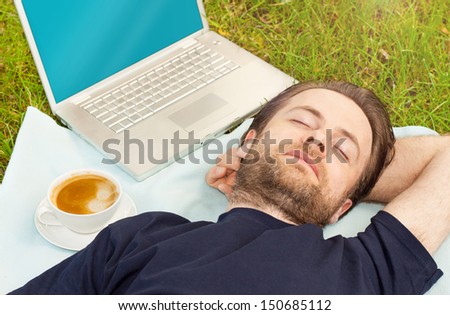 Man sleeping outdoor on grass next to a coffee and laptop computer - freelance work or exhausted designer concept