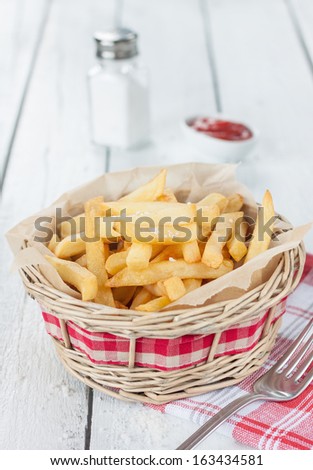 French fries in a wicker basket on white wood table with salt shaker and ketchup - rural bar or fast food menu
