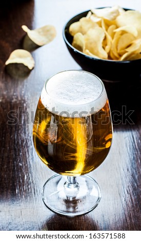 Beer glass and potato chips in a black bowl on dark wood background - snack bar or pub menu