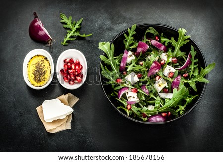 Fresh spring salad with rucola, feta cheese, red onion and pomegranate seeds in black bowl. Recipe ingredients on chalkboard background.