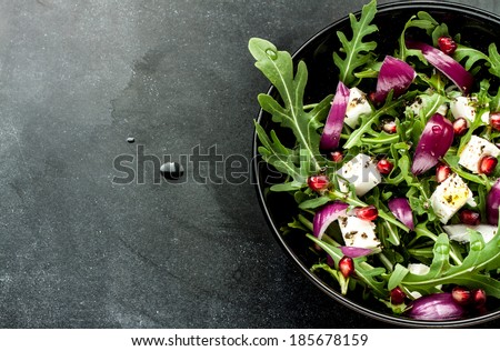 Fresh spring salad with rucola, feta cheese, red onion and pomegranate seeds in black bowl on chalkboard background with free text space.
