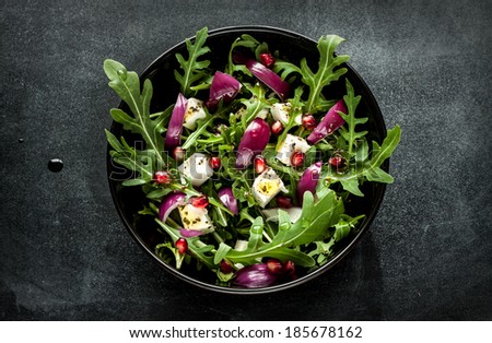 Fresh spring salad with rucola, feta cheese, red onion and pomegranate seeds in black bowl on chalkboard background.