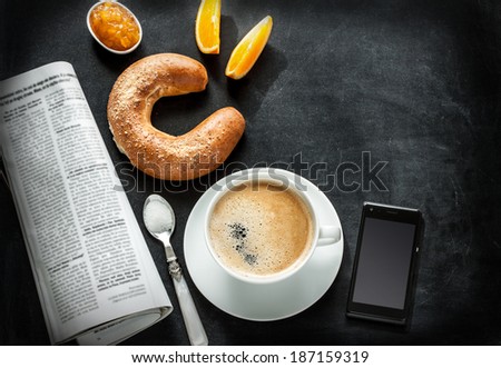 Continental breakfast on black chalkboard - bar menu. Coffee, orange juice, crescent roll, jam, newspaper and mobile phone from above. Background layout with free text space.