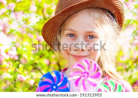 Portrait of happy smiling five years old caucasian blond child girl in a hat on blooming garden background. Spring - careless childhood, playing windmill.