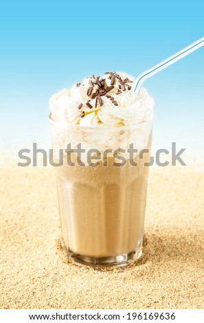 Summer iced coffee (frappuccino, frappe or latte) with whipped cream in a tall glass on sand background - beach bar holiday cold beverages menu. Layout with free text space.