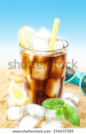 Cold cola or ice tea in a tall glass with lemon, mint leaves on sand background - beach bar summer holiday cold beverages menu. Layout with free text space.