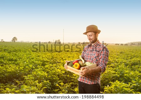 Happy smiling caucasian forty years old farmer in a hat standing proud in front of field landscape holding wooden box with fresh vegetables - agriculture.