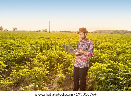 Happy smiling caucasian forty years old modern farmer in a hat standing outdoor in front of field landscape and working on laptop computer - agriculture. Layout with free text space.