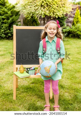 Happy smiling six years old blond caucasian child girl standing outdoor in front of the chalkboard - back to school, education concept.