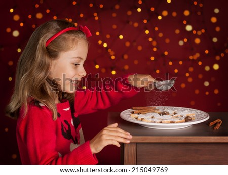 Christmas - happy smiling six years old blond caucasian child girl decorating cookies plate on dark red background with lights
