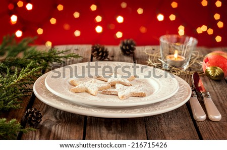 Christmas table - elegant white plate with cookies, natural pine tree branch and pinecones on vintage planked wood. Rural or rustic style table setting on red backdrop with blurred lights.