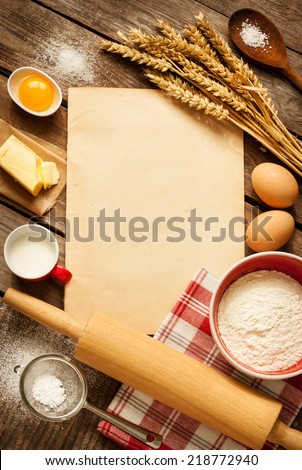 Rural vintage wooden kitchen table with old blank sheet of paper, baking cake ingredients (eggs, flour, milk, butter, sugar) and cooking utensils around. Background with free recipe text space.