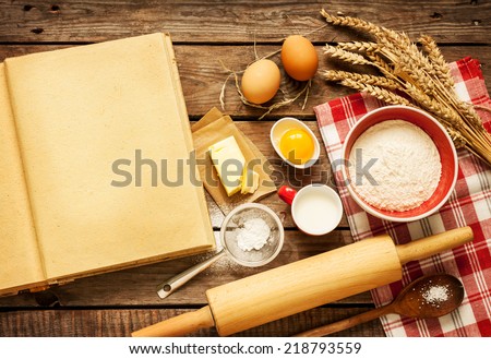 Rural vintage wooden kitchen table with blank cook book, baking cake or dough ingredients (eggs, flour, milk, butter, sugar) and cooking utensils around. Background with free recipe text space.