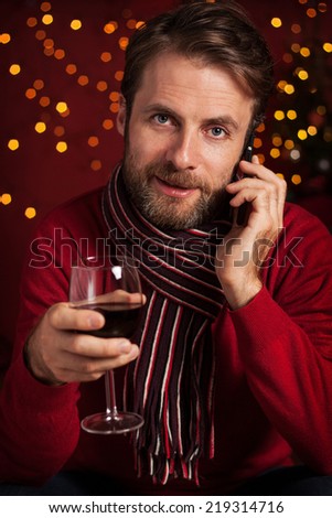 Christmas - happy smiling forty years old caucasian man with a glass of wine talking on mobile phone. Dark red with lights as background.