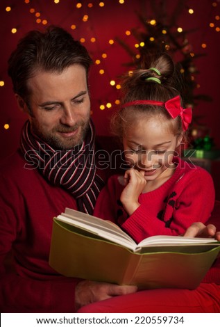 Christmas eve - happy family time. Smiling father and daughter read book on dark red background with lights.