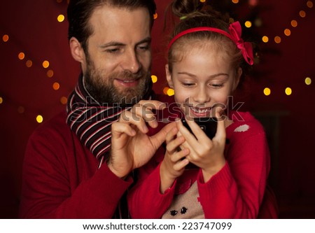 Christmas - smiling father and daughter playing game on mobile phone. Happy family time, dark red with lights as background.