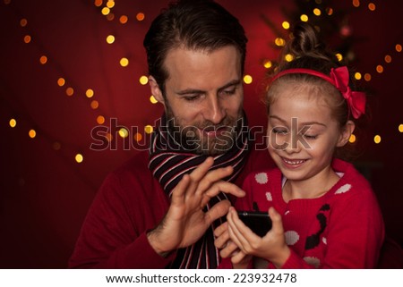 Christmas - smiling father and daughter playing game on mobile phone. Happy family time - modern lifestyle. Christmas tree with lights on dark red as background.