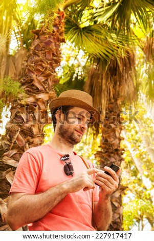 Happy smiling forty years old caucasian tourist man looking at mobile phone outdoor among exotic palm trees - summer holiday traveling