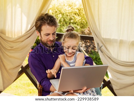 Smiling father and daughter playing game on laptop computer during sunny summer day. Happy family time  outdoor on garden terrace - modern lifestyle or holiday concept.