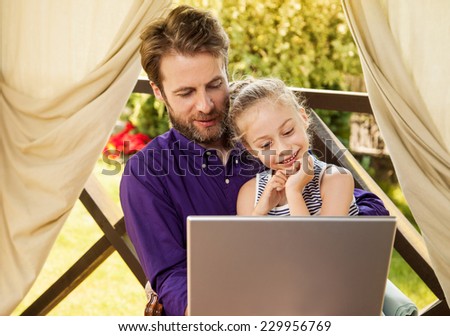 Smiling father and daughter looking at laptop computer during sunny summer day. Happy family time outdoor on garden terrace - modern lifestyle or holiday concept.