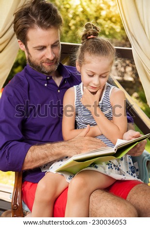Smiling father and daughter reading book during sunny summer day. Happy family time outdoor on garden terrace - countryside weekend or holiday concept.