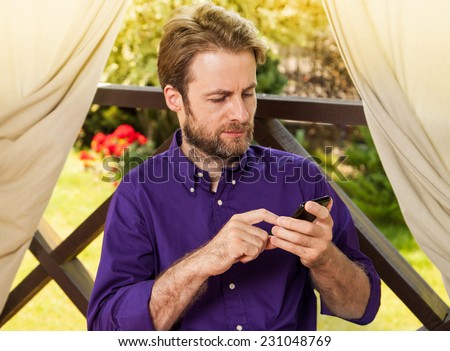 Forty years old caucasian man looking at mobile phone outdoor on garden terrace during sunny summer day. Countryside weekend or holiday concept.