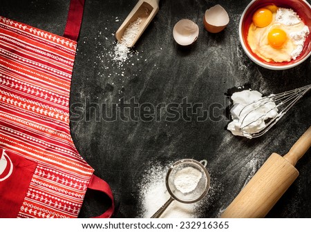 Baking cake ingredients. Apron, bowl, flour, eggs, egg whites foam, eggbeater, rolling pin and eggshells on black chalkboard from above. Cooking course poster background - layout with free text space.