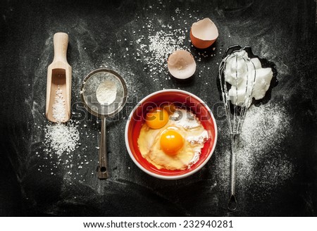 Baking cake ingredients - bowl, flour, eggs, egg whites foam, eggbeater and eggshells on black chalkboard from above. Cooking course or kitchen mess poster concept.
