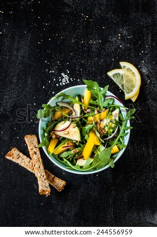 Fresh green spring salad with arugula (rucola), yellow pepper, zucchini, sunflower seeds and croutons on black chalkboard background from above. Poster layout with free text space.