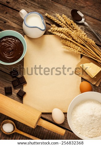 Rural vintage wooden kitchen table with old blank sheet of paper, baking cake ingredients (chocolate, eggs, flour, milk, butter, sugar) and cooking utensils around. Background layout.
