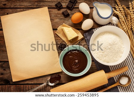 Rural vintage wooden kitchen table with old blank sheet of paper and baking cake ingredients (chocolate, eggs, flour, milk, butter, sugar). Background layout with free recipe text space.