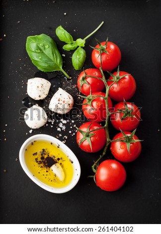 Cherry tomatoes, mozzarella cheese, basil and olive oil on black chalkboard from above. Italian caprese salad recipe ingredients.