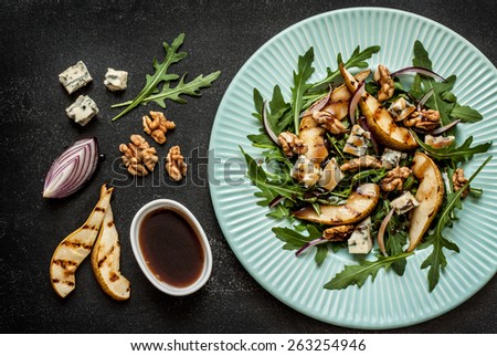 Salad recipe - blue cheese, pear, arugula, walnuts, red onion and balsamic vinegar dressing on pastel blue plate from above. Black chalkboard as background.