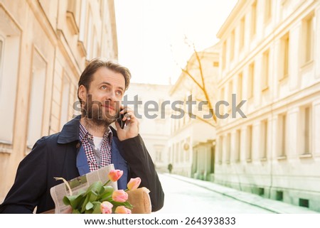 Happy smiling forty years old caucasian man with flower bouquet talking on a mobile phone. Street and city buildings as background.