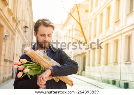 Forty years old caucasian man with baguette, newspaper and flower bouquet looking at watch. Street and city buildings as background.