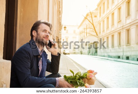 Happy smiling forty years old caucasian man with flower bouquet talking on a mobile phone. Street and city buildings as background.