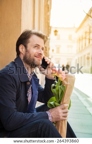 Happy smiling forty years old caucasian man with baguette and flower bouquet talking on a mobile phone. Street and city buildings as background.