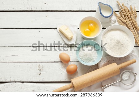Baking cake in rustic kitchen - dough recipe ingredients (eggs, flour, milk, butter, sugar) on white planked wooden table from above. Background layout with free text space.