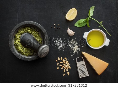 Green basil pesto - italian recipe ingredients on black chalkboard from above. Parmesan cheese, basil leaves, pine nuts, olive oil, garlic, salt, pepper and mortar.