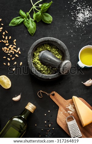 Green basil pesto - italian recipe ingredients on black chalkboard from above. Parmesan cheese, basil leaves, pine nuts, olive oil, garlic, salt, pepper and mortar.