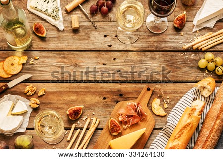 Different kinds of cheeses, wine, baguettes, fruits and snacks on rustic wooden table from above. French tasting party or feast scenery. Layout with free text space.