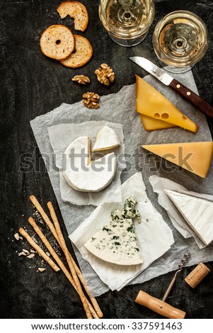 Different kinds of cheeses, white wine and snacks on black chalkboard background captured from above (top view). French tasting party or feast scenery.