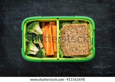 Green school lunch box with sandwich, broccoli and carrot close up on black chalkboard background. Healthy eating habits concept. Flat lay composition (from above, top view).