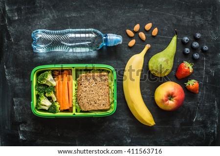School lunch box with sandwich, vegetables, water, almonds and fruits on black chalkboard background. Healthy eating habits concept. Flat lay composition (from above, top view).