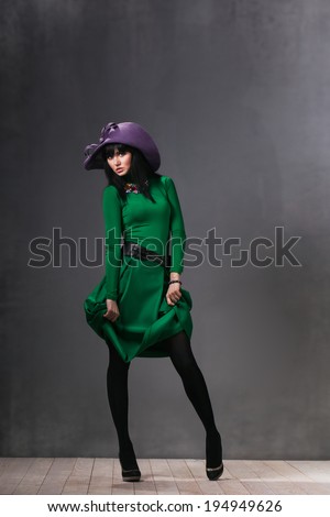 Beautiful sexy woman in a green dress and purple hat posing in the studio and slightly lifts the dress hands