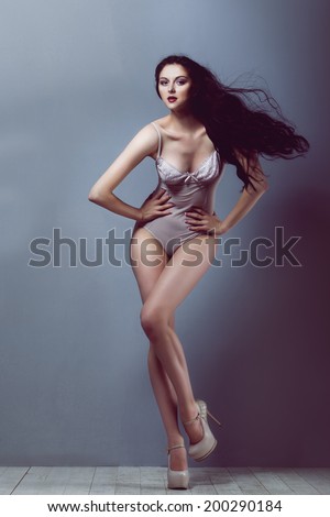 sexy brunette girl in a transparent body beige heels with a beautiful body and long legs and hair developing on a wind in studio on white wooden floor and gray walls
